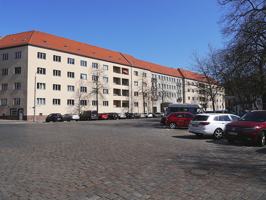 Sehr breite gepflasterte Straße, dahinter Wohnhaus, Parkraum