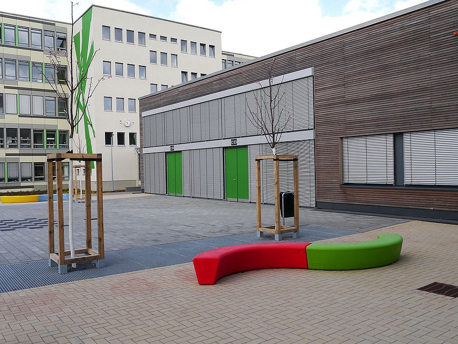 Schulgebäude, flacherer Verbindungsbau, Jungbäume, Sitzschlange in rot-grün