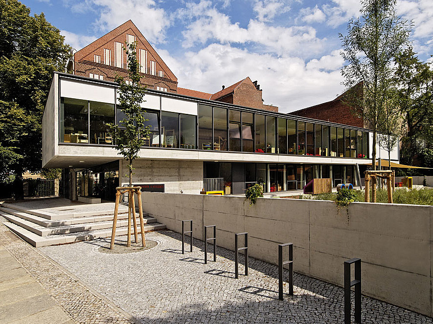 Moderner Zweigeschosser mit breiterem Obergeschoss und durchgehender Fensterfront, dahinter Backsteingiebel, Park mit neu gepflanzten Bäumen