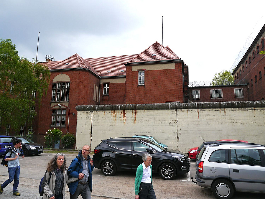 Parkende Autos und Menschen vor Mauer, dahinter Backsteingebäude