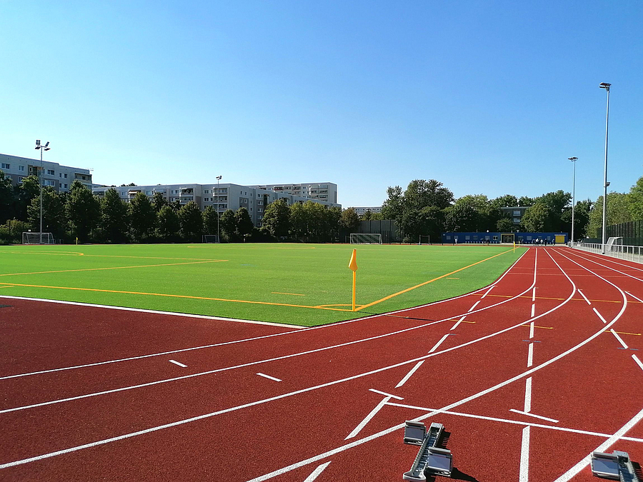Blick von Startblöcken auf gelbe Eckfahne und grünes Fußballfeld, im Hintergund Wohnhäuser