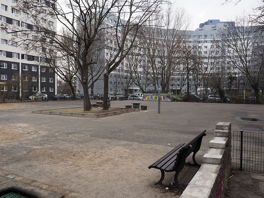 Platz mit Baum im Winter in der Mitte, am Rand Gebäuderiegel