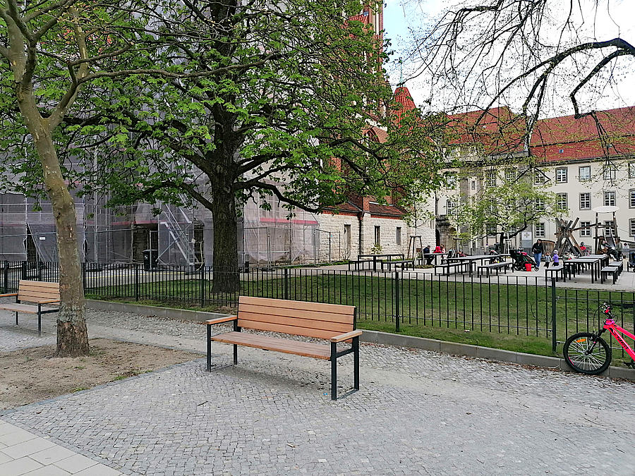 Holzbank, dahinter niedrig eingezäunte Grünfläche, Picknicktische, Kirche