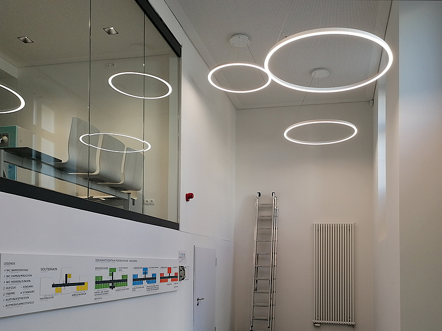 Im Eingangsbereich schafft ein Wand- und Deckendurchbruch mehr Licht  und Offenheit Hoher, schmaler Raum mit kreisförmigen Deckenleuchten und Durchblick zum Wartebereich, Infotafel