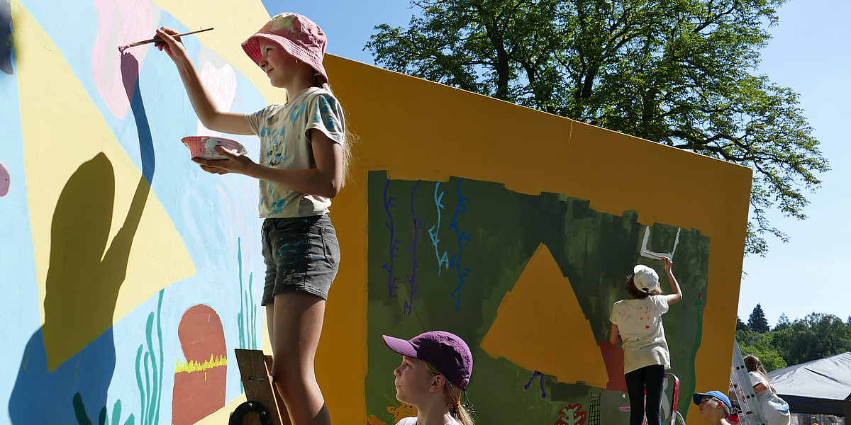 Kinder auf Leitern malen an einer hohen, geknickten, bunten Wand
