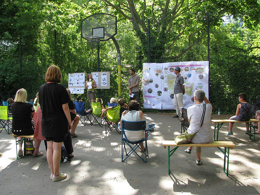 Kinder und Erwachsene auf Bänken oder stehend vor Plakaten am Zaun, ein Mann erklärt