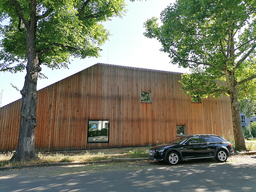 Holzlatten-Fassade eines zweistoeckigen Gebaeudes mit asymmetrischen Fenstern, davor Straße mit Bäumen und parkendem PKW