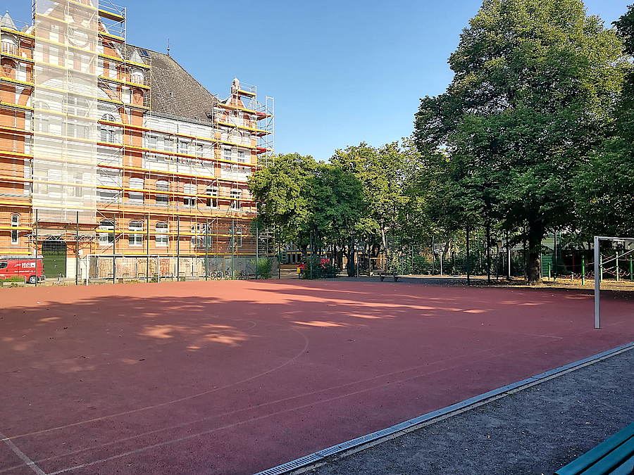 Historisches Schulgebäude eingerüstet, davor Sportplatz (Hartplatz) mit Fußballtor