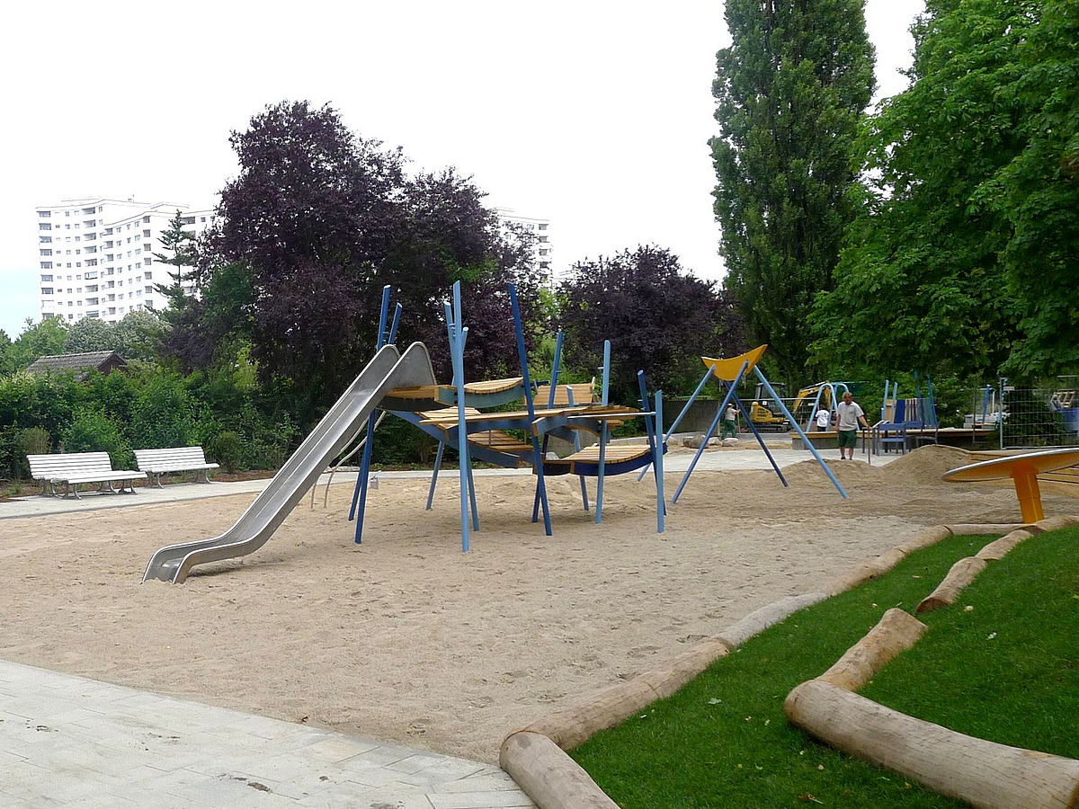 Sandspielplatz mit Rutsche umgeben von Bäumen, im Hintergrund Hochhäuser