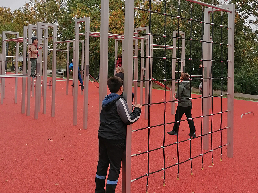 Kinder an senkrechtem Kletternetz auf rotem Belag im Park