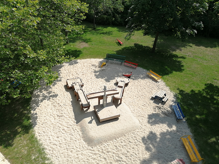 Vogelperspektive auf ovale Sandfläche mit Rinnen-Anlage und Bänken, ringsum Rasen, große Bäume