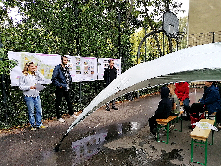 2 Menschen vor Plakaten am Zaun, Kinder und andere Gäste unter Regenschutz auf Bänken