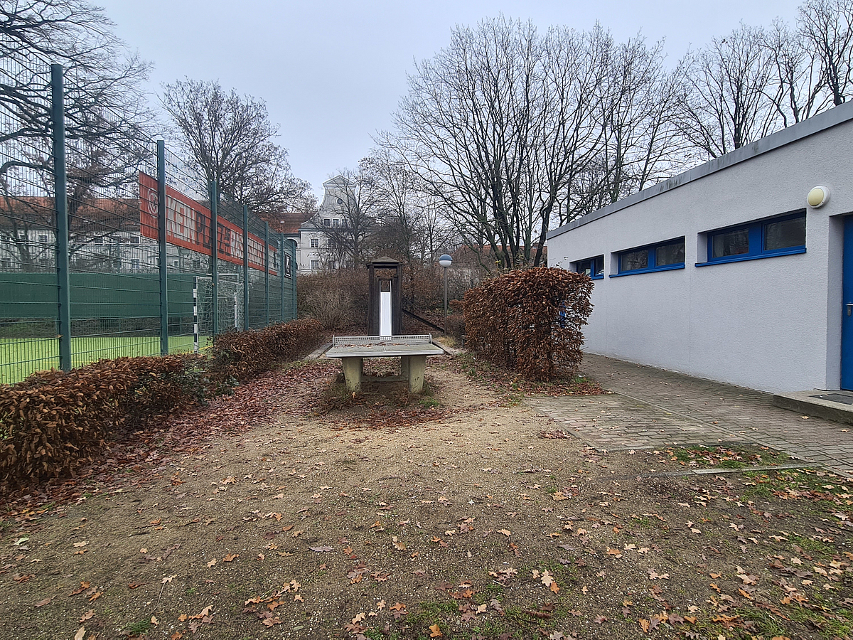 Kleiner, eingezäunter Bolzplatz, Funktionsgebäude, dazwischen Tischtennisplatte und Rutsche