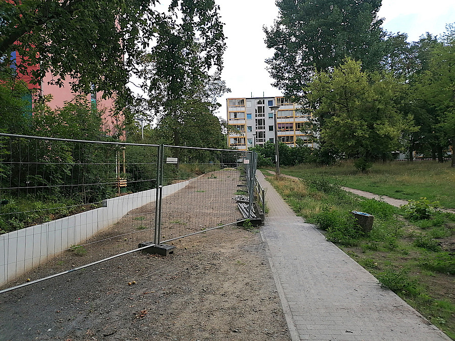 Bauzaun, daneben schmale Rampe, dahinter Wohnblock