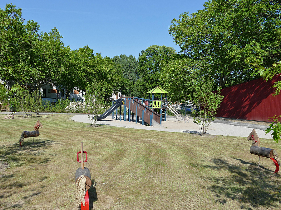 Kitagarten mit Rasen, Holzpferden und Sandspielplatz mit Kletterturm und Rutsche, Grün, dunkelrote Holzwand