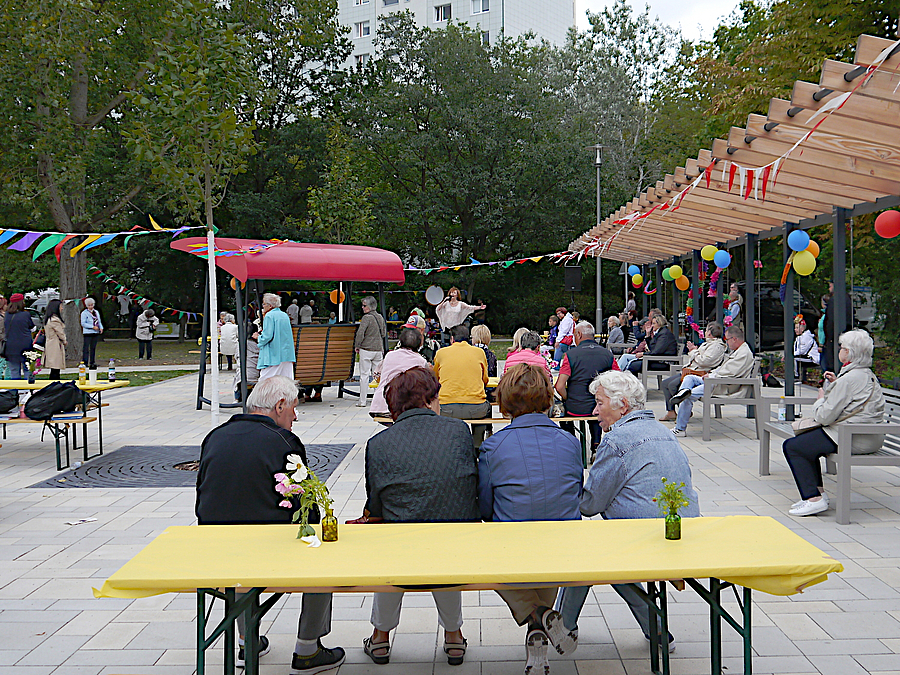Menschen sitzen auf Platz auf Bänken unter Pergola, im Hintergrund Grün