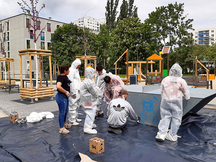 Kinder aus der Gemeinschaftsunterkunft und einem Jugendclub bei der gemeinsamen Arbeit mit Sozialarbeiter Jan Blüm 4 Personen in weißen Schutzanzügen, 1 Person in normaler Kleidung an trapezförmigem Körper auf Folie im Freien, im Hintergrund blühende junge Bäume, Spielplatz