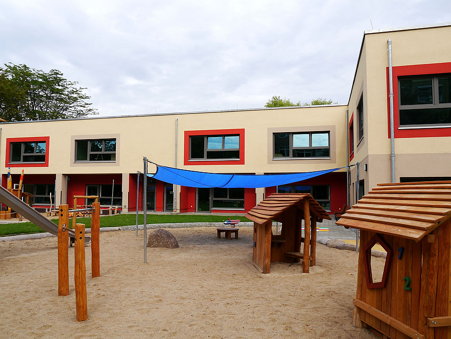 Gebäude in L-Form, Sandspielfläche, Spielhütten, Sonnensegel