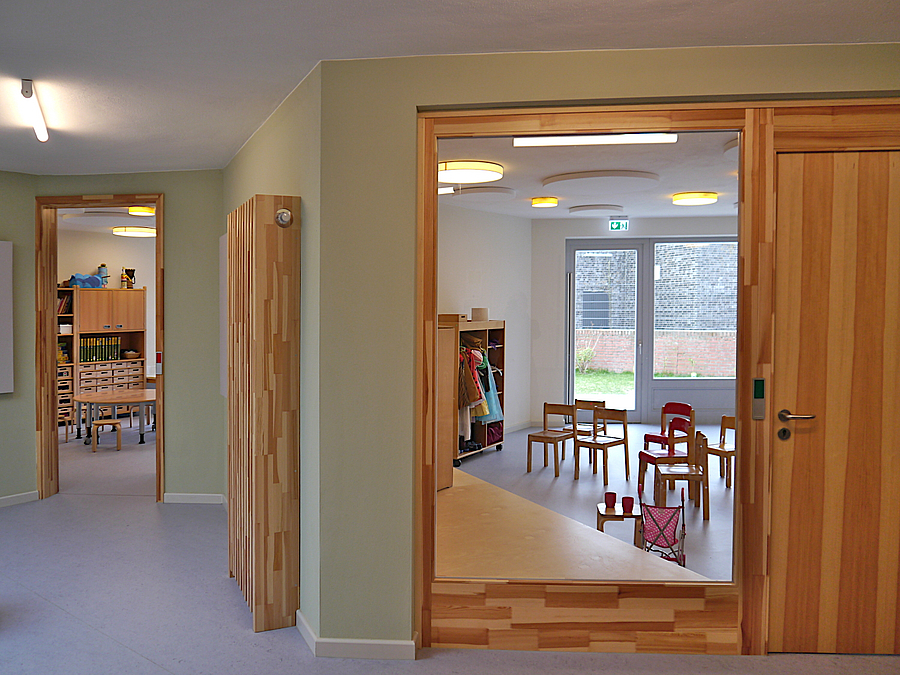 Flur in pastellgrün, holzverkleidete Heizung, Holztür und großes Fenster zum Gruppenraum