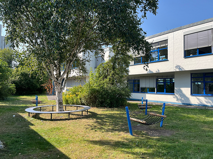 Heller Zweigeschosser hinter Wiese mit Birke und Rundbank