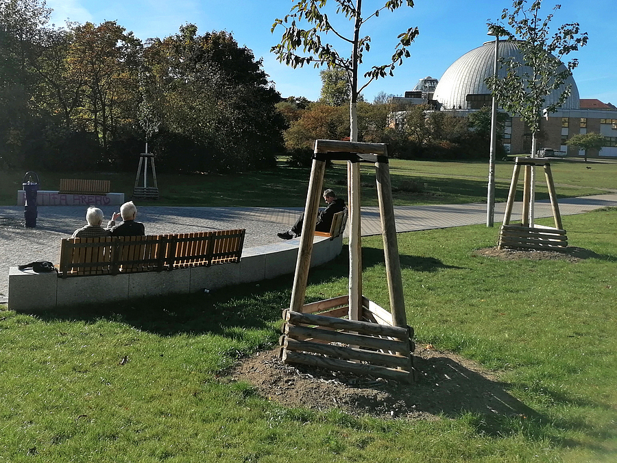 Jugen Bäume und besetzte Bänke im Park, im Hintergrund Planetarium