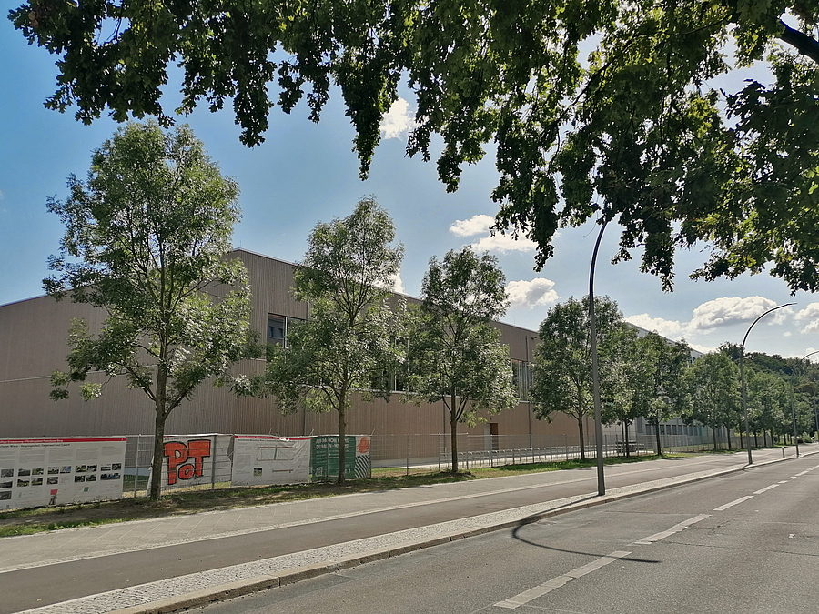 Straße mit Geh- und Radweg sowie Holzmodul-Sporthalle und Infoplakat am Bauzaun