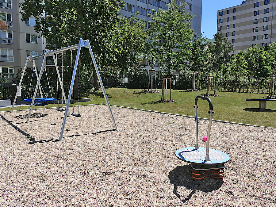 Kiesfläche mit Schaukeln und Drehwippe, dahinter Rasen mit jungen Bäumen