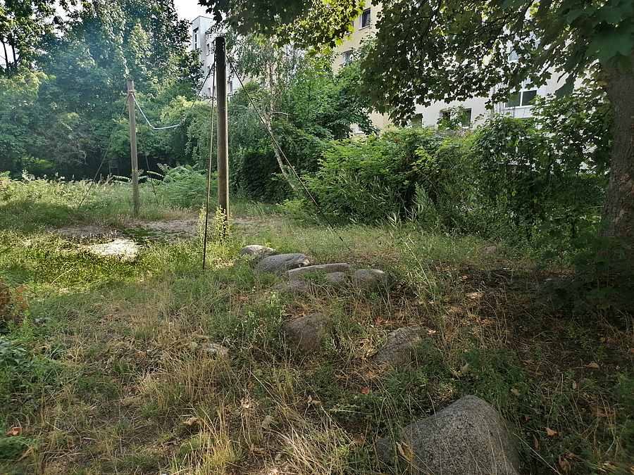 Zuwachsende Grünfläche mit kleinen Findlingen und Holzmasten mit Seil ohne Funktion