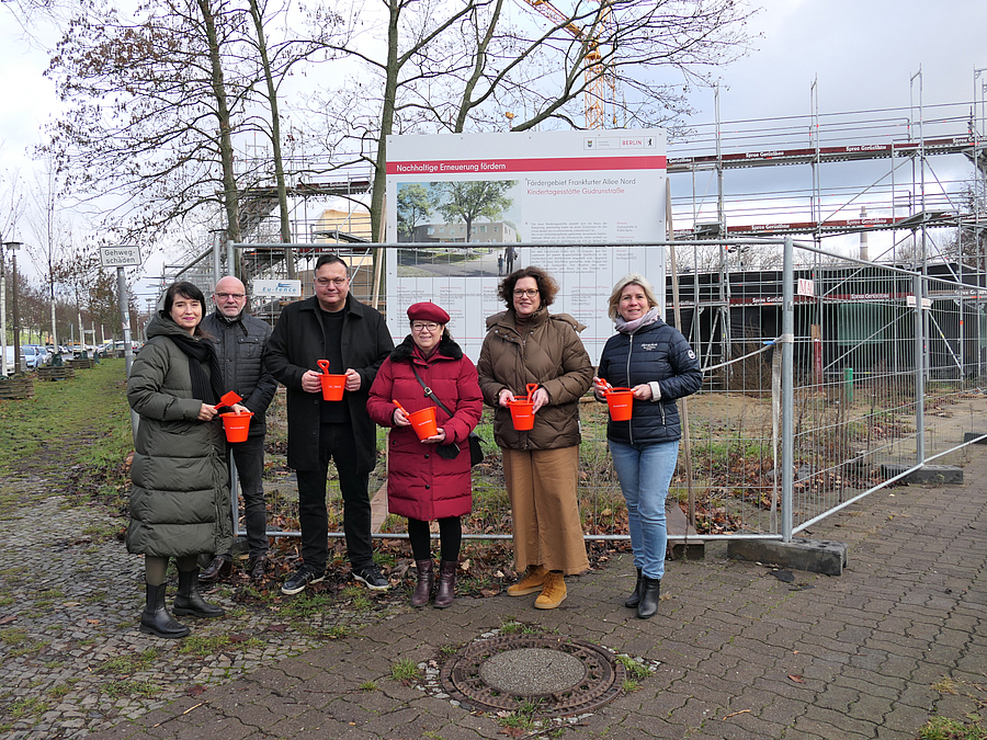6 Personen mit Buddeleimern vor Bauschild und Baustelle