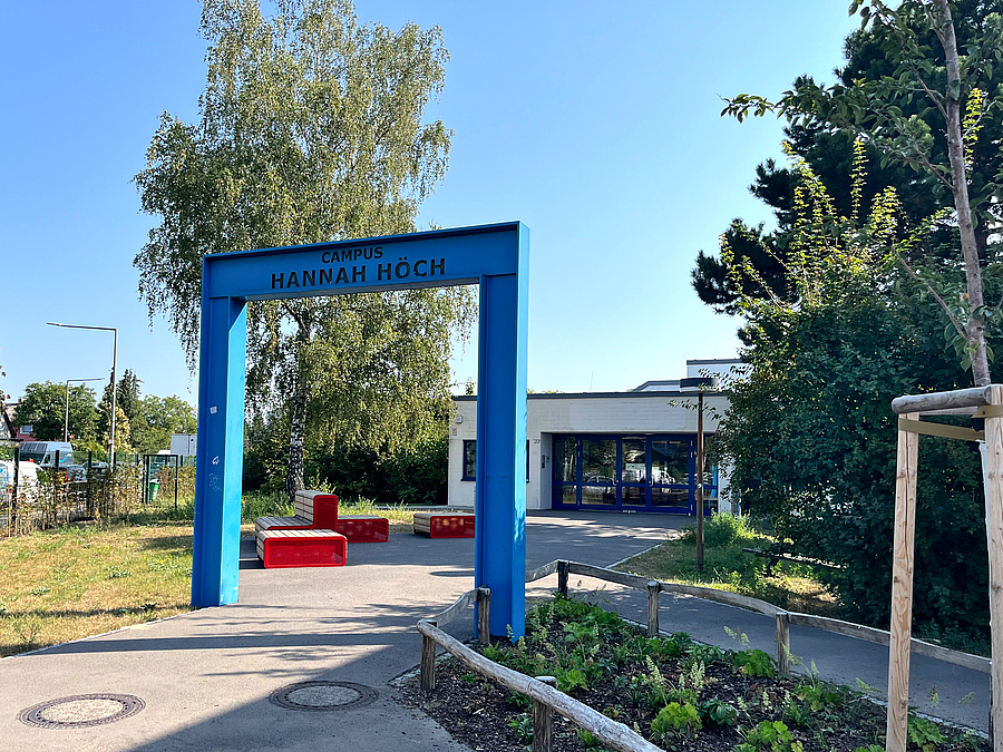 Der Eingang zum Oberschulteil des Campus mit neuen Möbeln und Pflanzungen Rechteckiger, blauer Stahlrahmen mit Schulname, Weg, Beet, Rote Außenmöbel, Bäume