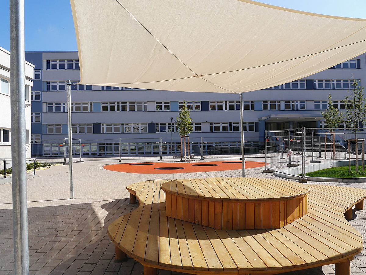 Zweistufiges Holzdeck mit Sonnensegel, junge Bäume, Plattenbau-Schule