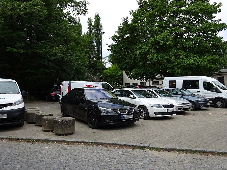 PKW und Lieferwagen parken auf baumgesäumtem Platz, dazwischen eine Reihe Betonzylinder