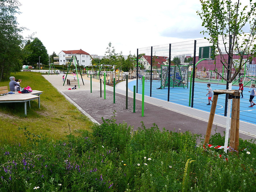 Im Vordergrund Blühwiese, Rasen mit besetztem Holzdeck, blaues Ballspielfeld, Seilbahn
