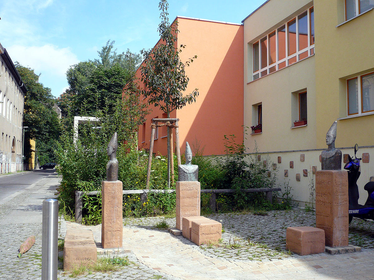 Gebäude mit Vorsprung in Gelb und Orange, Stelen mit Büsten von Harlekins auf Pflasterfläche davor
