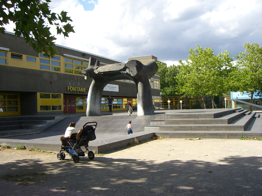 Platz mit Brunnen im brutalistischen Stil, Frau mit Kinderwagen, grauer Flachbau mit gelben Fensterrahmen